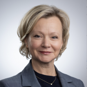 Headshot of a person with short, light-colored hair, wearing a blue blazer and a dark top, set against a neutral background.